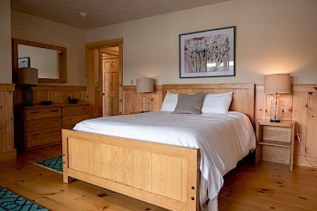 A bright cabin bedroom with light wood paneling and hardwood floors, featuring a wooden bed frame, dresser, and modern art.
