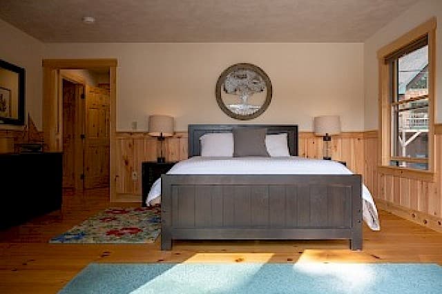 A bright cabin bedroom with a dark wood bed and light wood paneling, featuring a round mirror and two matching bedside tables and lamps.