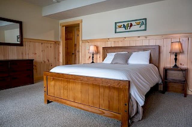 A cozy cabin bedroom with natural wood paneling, a light wood bed frame, two lamps, a dark wood dresser, and a carpeted floor.