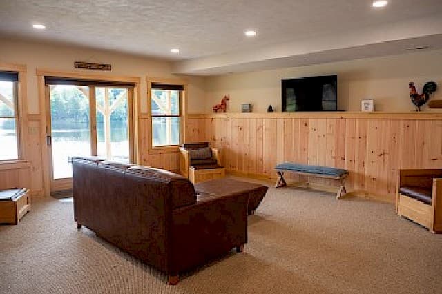A lower-level living room with a brown leather sofa, wood paneling, a flat-screen TV, and a glass door opening to an outdoor area.