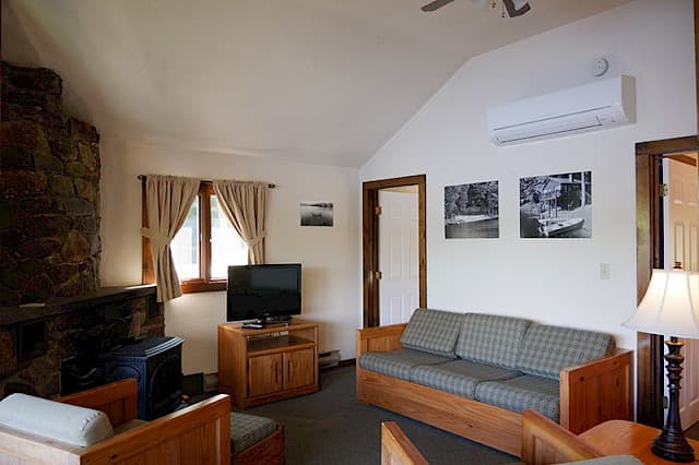 A cozy cabin living room with a vaulted ceiling, a large stone fireplace, a wooden futon/sofa, and a flat-screen TV.
