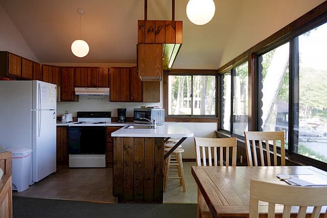 A bright cabin kitchen and dining area with a vaulted ceiling, wood cabinets, a white fridge, and a dining table next to large windows.