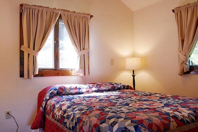 A cozy bedroom with a vaulted ceiling, a bed with a colorful patterned quilt, a bedside lamp, and two curtained windows.
