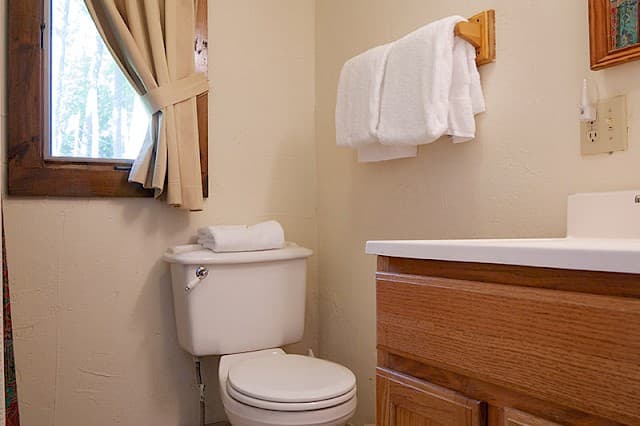 A simple, well-lit bathroom with a white toilet, wooden vanity, towels on a wooden rack, and a small, curtained window.