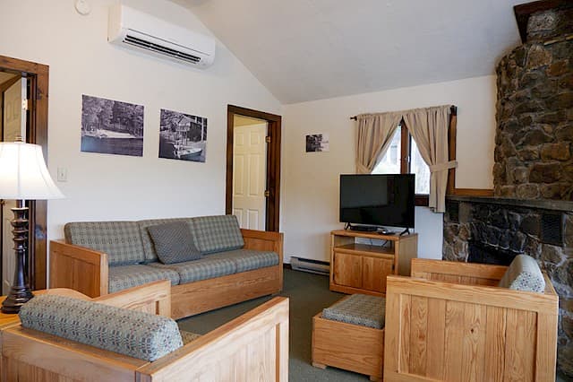 A cozy cabin living room with a vaulted ceiling, natural wood furniture, a stone fireplace on the right, and a small TV on a stand.