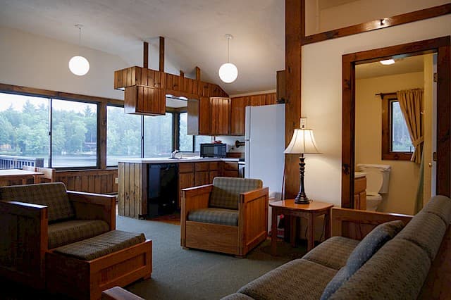 An open-concept cabin space with a vaulted ceiling, featuring a living area, kitchen with wood cabinets, and large windows overlooking a lake.