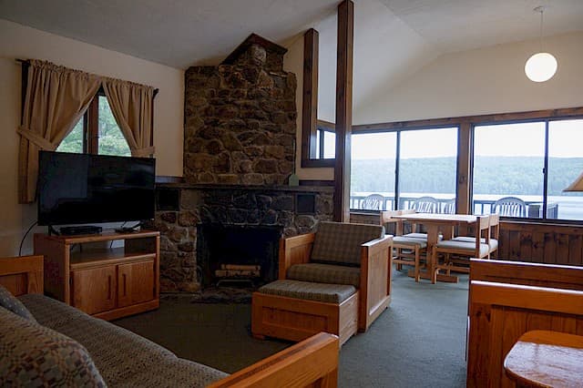 A rustic cabin living room with a vaulted ceiling, natural wood furniture, a large stone fireplace, and a wall of windows with a view.