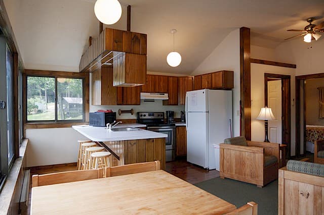 A cabin kitchen and dining area with a vaulted ceiling, wood cabinets, a white fridge, and a large dining table by a wall of windows.