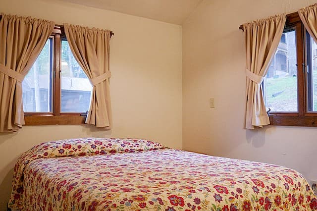 A simple cabin bedroom with a vaulted ceiling, a bed with a colorful floral comforter, and two windows with tied-back beige curtains.