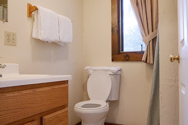 A simple, well-lit bathroom with a white toilet, wooden vanity, towels on a wooden rack, and a small window with beige curtains.
