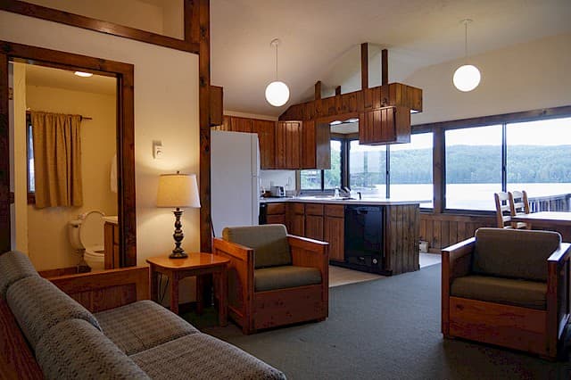 An open-concept cabin space with a vaulted ceiling, featuring a living area, kitchen with wood cabinets, and large windows overlooking a lake.