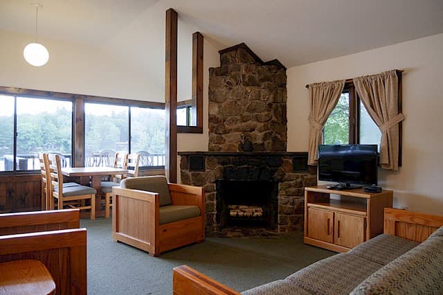 A rustic cabin living and dining area with a vaulted ceiling, a large stone fireplace, wood furniture, and a view through large windows.