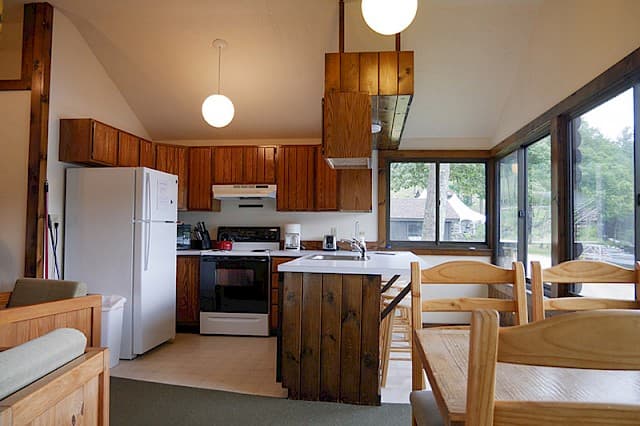 A bright cabin kitchen and dining area with a vaulted ceiling, wood cabinets, a white fridge, and a dining nook next to large windows.