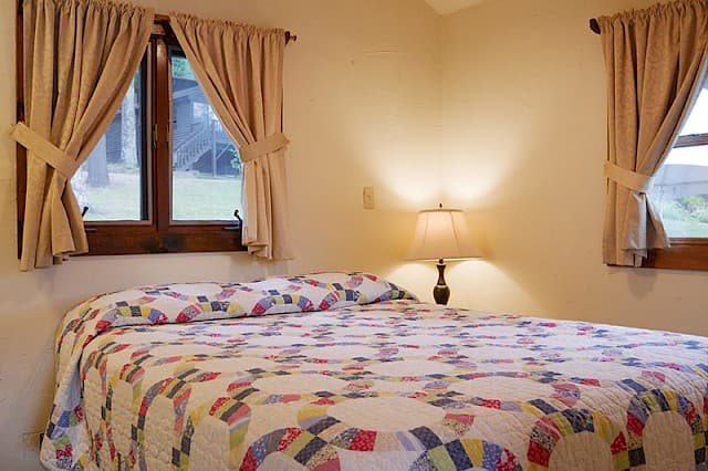 A cozy cabin bedroom with a vaulted ceiling, a bed with a ring-patterned quilt, and two windows with tied-back beige curtains.