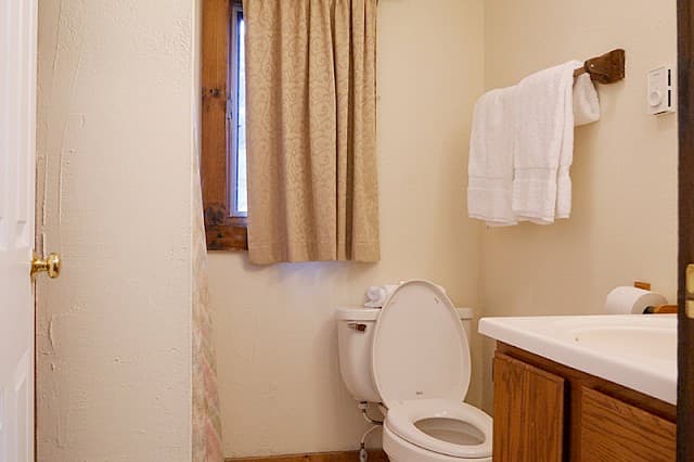 A simple, well-lit bathroom with a white toilet, wooden vanity, towels on a wooden rack, and a small, curtained window.
