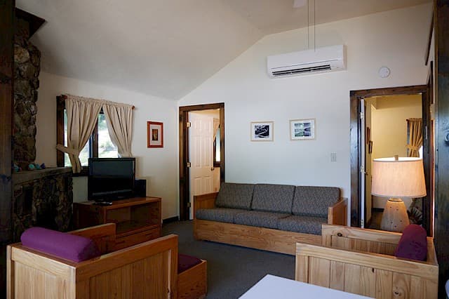 A cozy cabin living room with a vaulted ceiling, natural wood furniture and a sofa, featuring a large stone fireplace and a mini-split AC unit.