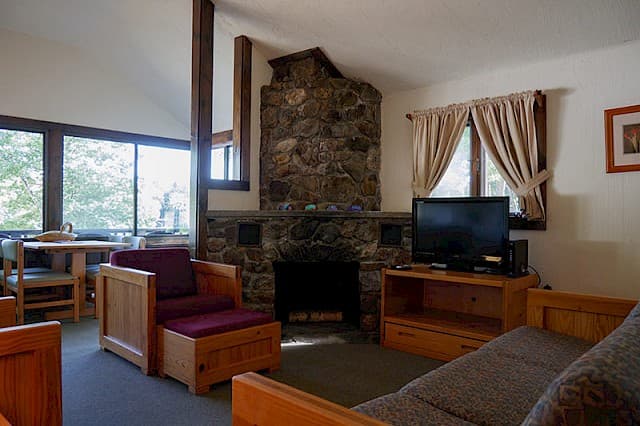 A rustic cabin living and dining area with a vaulted ceiling, a large stone fireplace, wood-framed furniture, and large windows with a view.