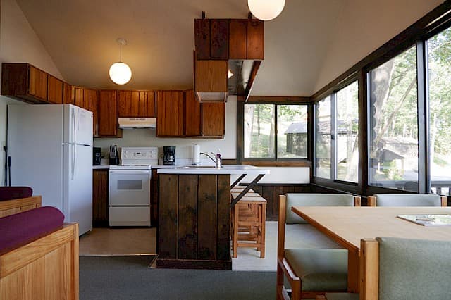 A cabin kitchen and dining area with a vaulted ceiling, wood cabinets, white appliances, and a dining nook next to large windows.