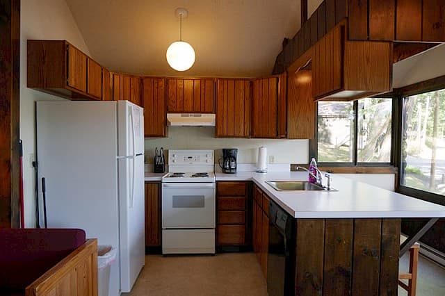 A cabin kitchen with a vaulted ceiling, wood cabinets, white appliances, and a breakfast bar with wood paneling below.
