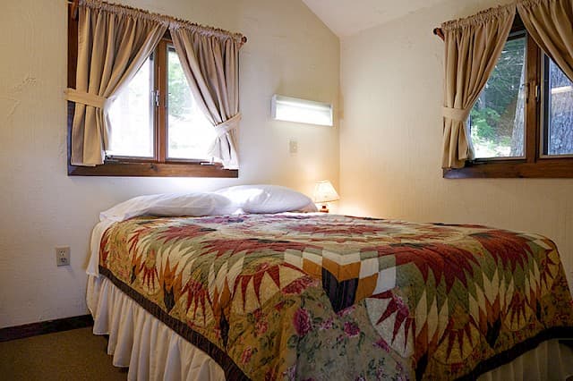 A cozy cabin bedroom with a vaulted ceiling, a bed featuring a colorful geometric quilt, and two windows with tied-back beige curtains.