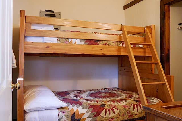 A simple cabin bedroom featuring a natural wood bunk bed with a ladder, and the lower bunk covered by a colorful patterned quilt.