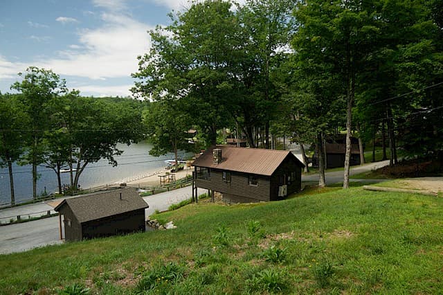 A view of a lake shore with two dark brown cabins, one elevated, and a grassy hill in the foreground under a bright, partially cloudy sky.