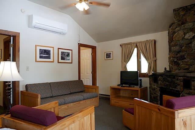 A cozy cabin living room with a vaulted ceiling, natural wood furniture, a large stone fireplace, and a mini-split AC unit.