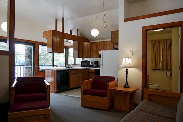 An open-plan cabin space with a vaulted ceiling, featuring a living area, kitchen with wood cabinets, and large windows leading to a deck.