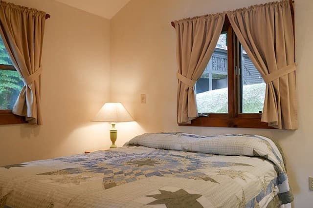 A simple cabin bedroom featuring a bed with a quilt, a bedside lamp, and two windows with tied-back beige curtains.