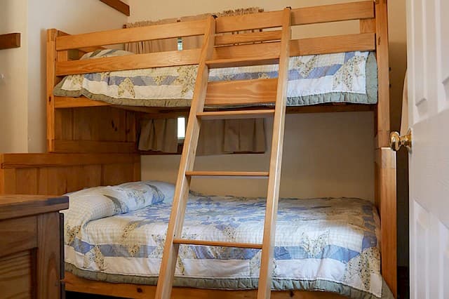 A simple cabin bedroom featuring a natural wood bunk bed with a ladder, both bunks covered by light blue and white patterned quilts.