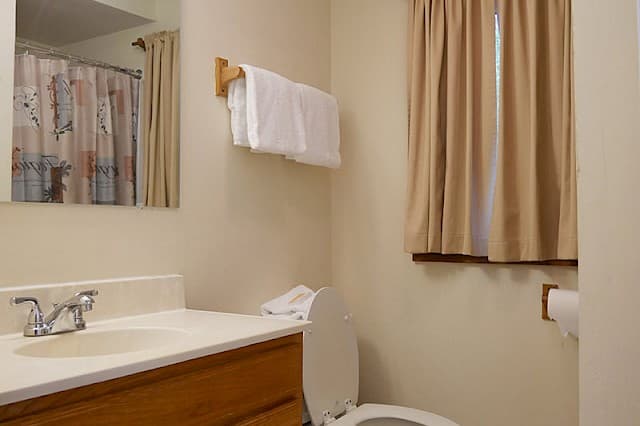 A simple cabin bathroom with a wooden vanity, white sink, a toilet, a towel rack with white towels, and a small curtained window.
