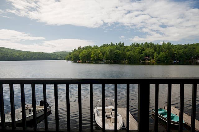 A wide view of a calm lake with a forested shoreline, seen from behind a dark railing, with a few small boats docked below.