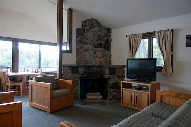 A rustic cabin living and dining area with a vaulted ceiling, a large stone fireplace, wood furniture, and a view through large windows.