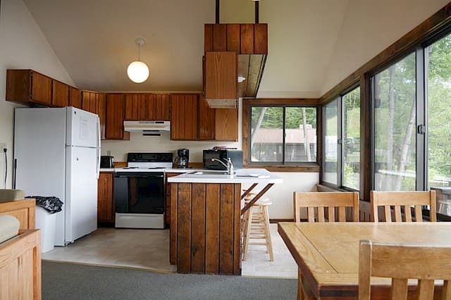 A bright cabin kitchen and dining area with a vaulted ceiling, wood cabinets, white appliances, and a dining table next to large windows.