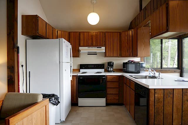 A cabin kitchen with a vaulted ceiling, wood paneling and cabinets, a white fridge, a black stove, and a single globe pendant light.