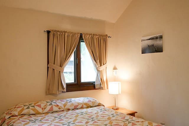 A simple cabin bedroom with a vaulted ceiling, a bed with a geometric patterned quilt, a bedside lamp, and a curtained window.