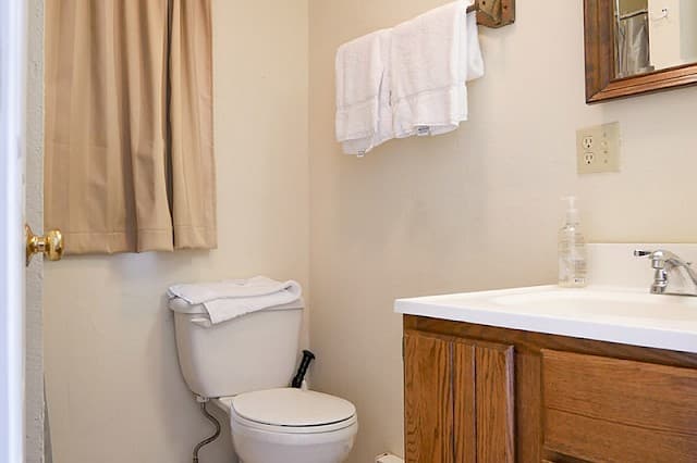 A simple cabin bathroom with a white toilet, wooden vanity, a towel rack with white towels, and a small, curtained window.