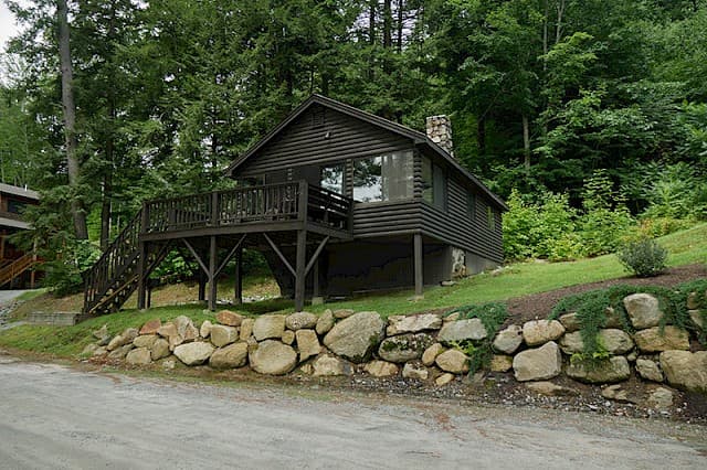 A dark brown elevated cabin with a deck and screened-in porch, set against a rock retaining wall and lush green hillside.