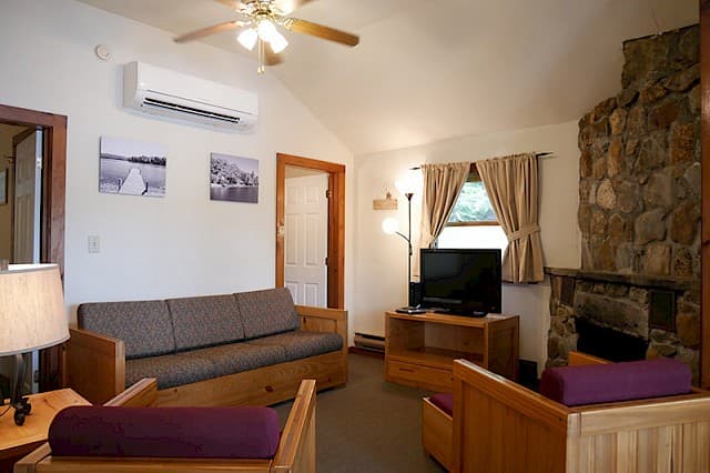 A cozy cabin living room with a vaulted ceiling, natural wood furniture, a large stone fireplace, and a mini-split AC unit.