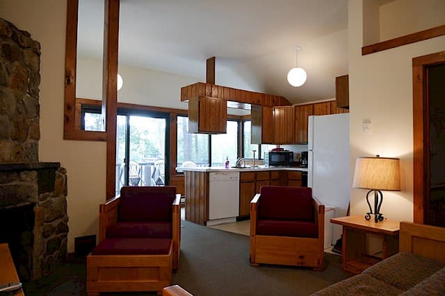 An open-plan cabin space with a vaulted ceiling, featuring a living area, kitchen with wood cabinets, and a stone fireplace on the left.