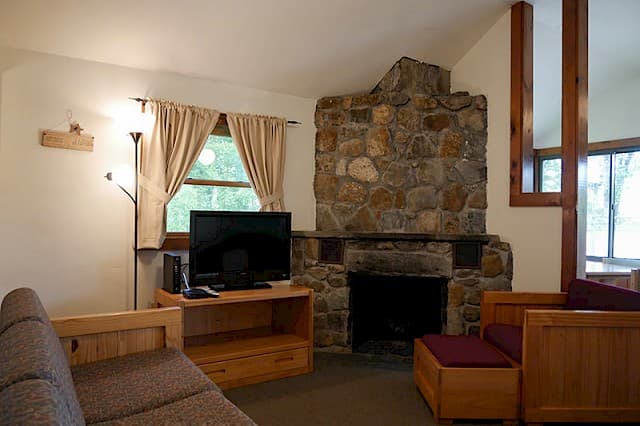 A cozy cabin living room with a vaulted ceiling, a large stone fireplace, wood furniture, and a small TV next to a curtained window.