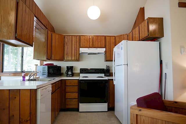 A cabin kitchen with a vaulted ceiling, wood paneling and cabinets, a white fridge, a black stove, and a single globe pendant light.