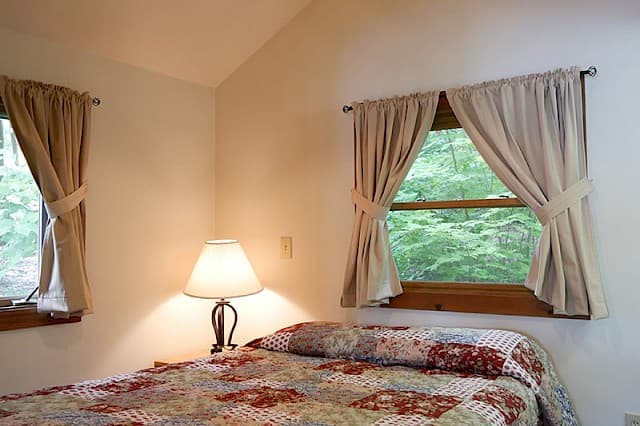 A simple cabin bedroom with a vaulted ceiling, a bed with a patterned quilt, a bedside lamp, and two curtained windows with a view of trees.