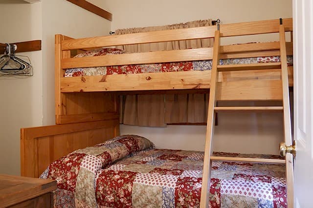 A simple cabin bedroom featuring a natural wood bunk bed with a ladder, and a quilt with a red and patterned patchwork design.