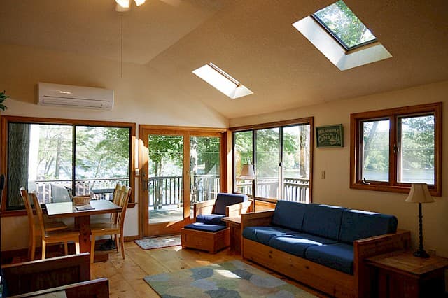 A bright cabin living area with a vaulted ceiling, skylights, a blue futon, a dining table, and large glass doors leading outdoors.
