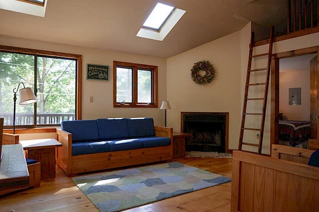 A bright cabin living room with a skylight, hardwood floors, a blue futon, a small fireplace, and a ladder leading to a loft area.