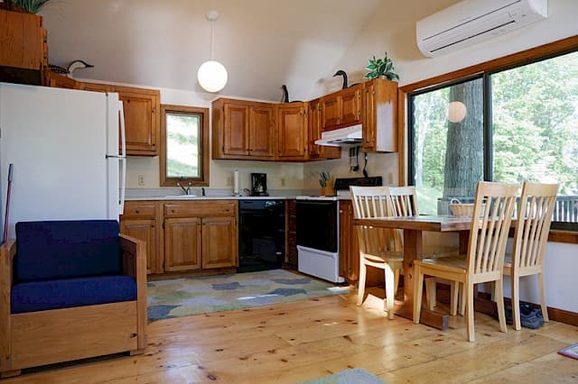 A bright cabin kitchen and dining area with a vaulted ceiling, hardwood floors, wood cabinets, a white fridge, and a wooden dining set.