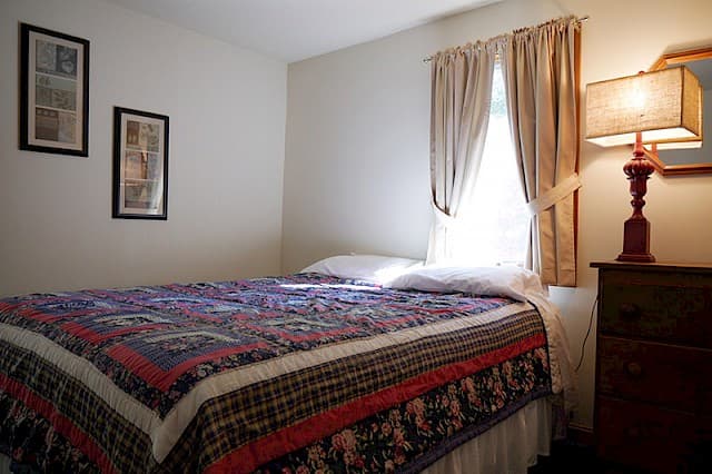 A cozy bedroom featuring a double bed with a colorful patterned quilt, a bedside table with a lamp, and a single window with beige curtains.
