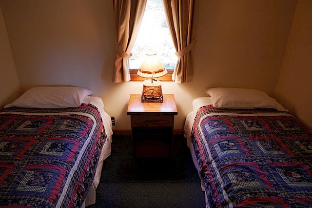 A simple cabin bedroom featuring two twin beds with patterned covers, separated by a wooden nightstand with a lamp under a curtained window.