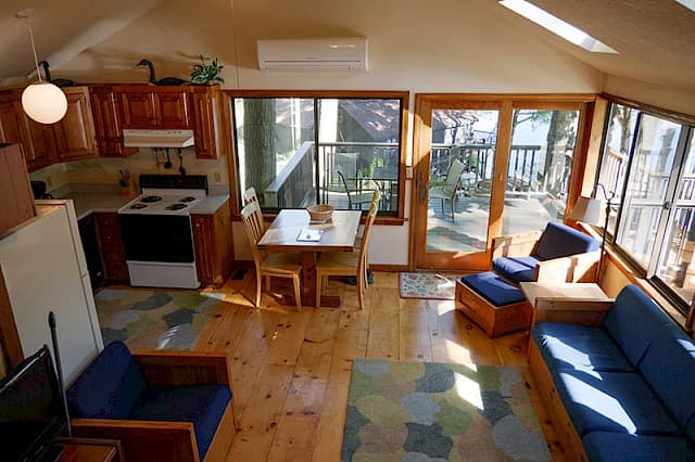 A bright cabin great room with a vaulted ceiling, hardwood floors, an open kitchen, and a sunlit living area with glass doors.